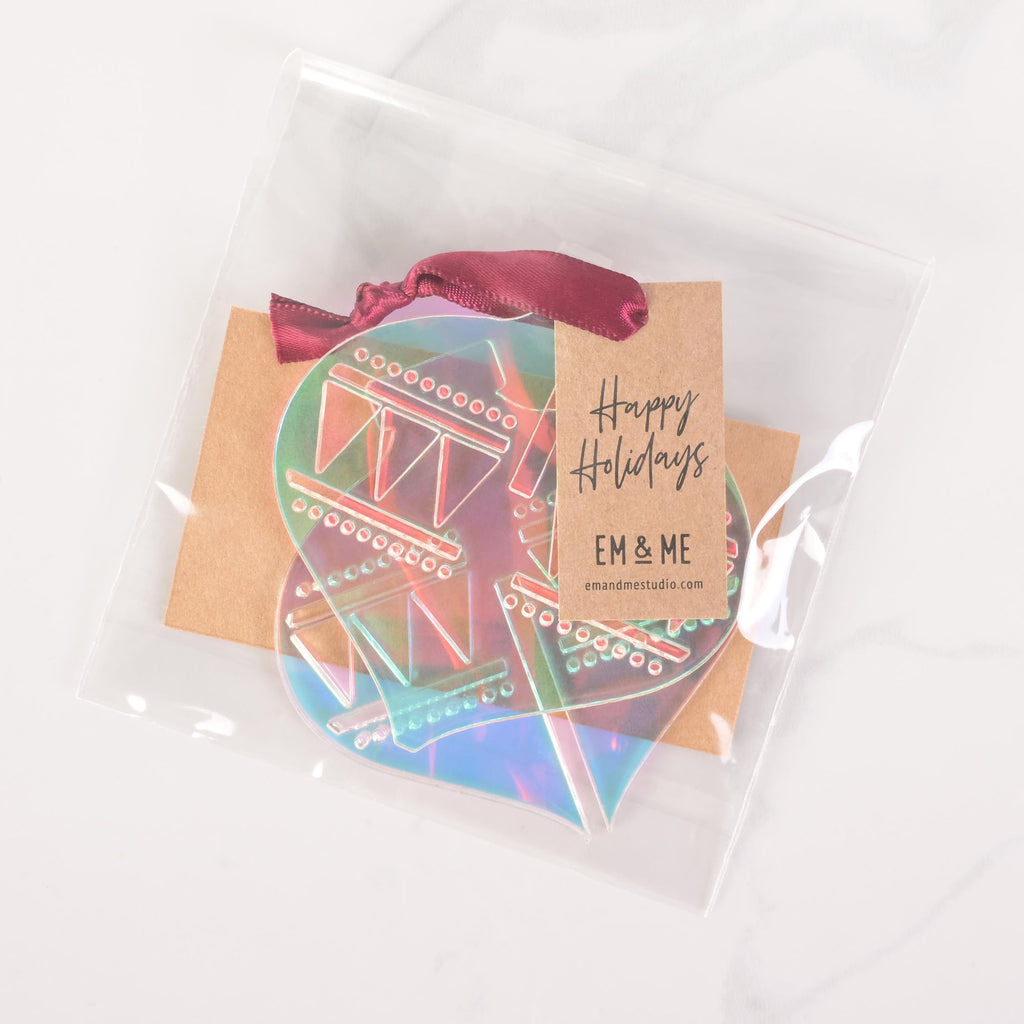 A packaged set of iridescent  flat-pack holiday ornament pieces. The clear plastic bag contains the parts needed to assemble an ornament and a brown cardstock tag that says "Happy Holidays" and "EM & ME." A dark ribbon is tied to the top of the package. The item is displayed on a white marble surface.