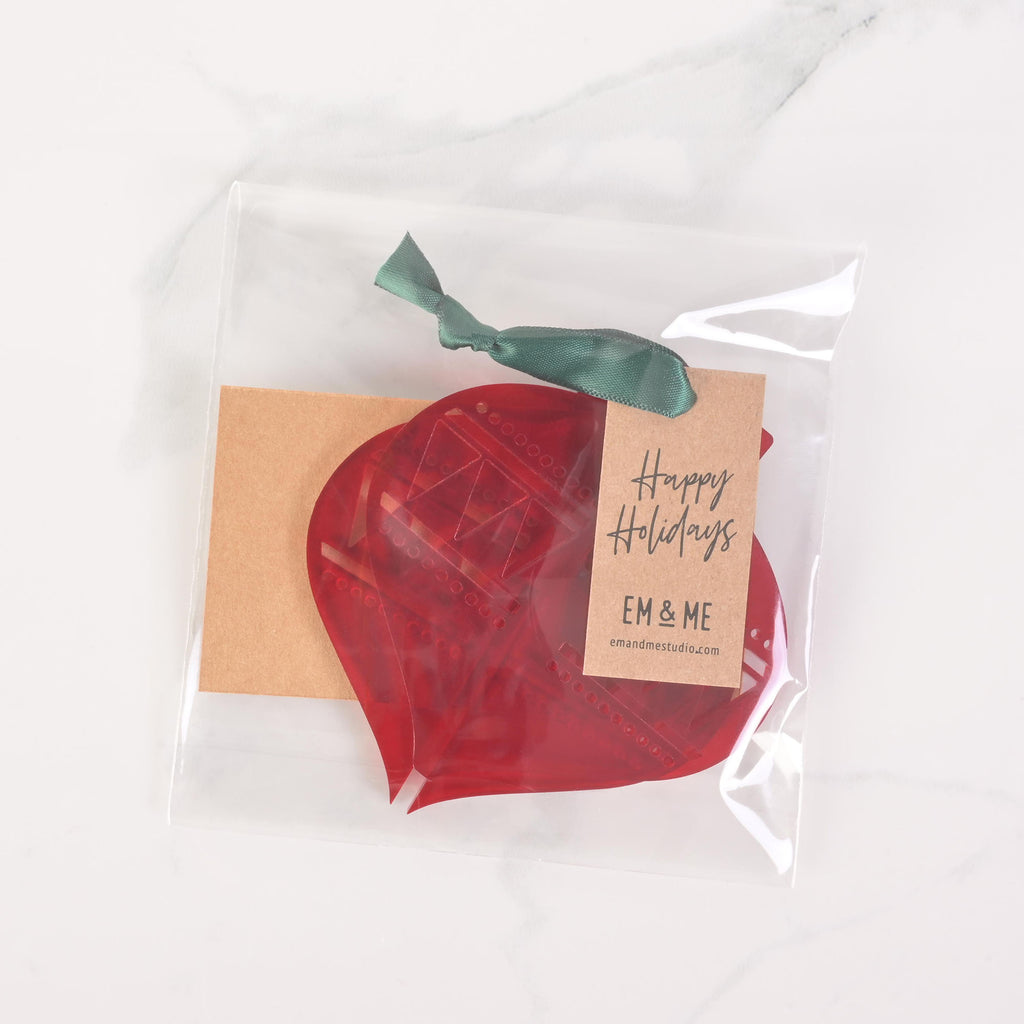 A packaged set of red flat-pack holiday ornament pieces. The clear plastic bag contains the parts needed to assemble an ornament and a brown cardstock tag that says "Happy Holidays" and "EM & ME." A dark ribbon is tied to the top of the package. The item is displayed on a white marble surface.