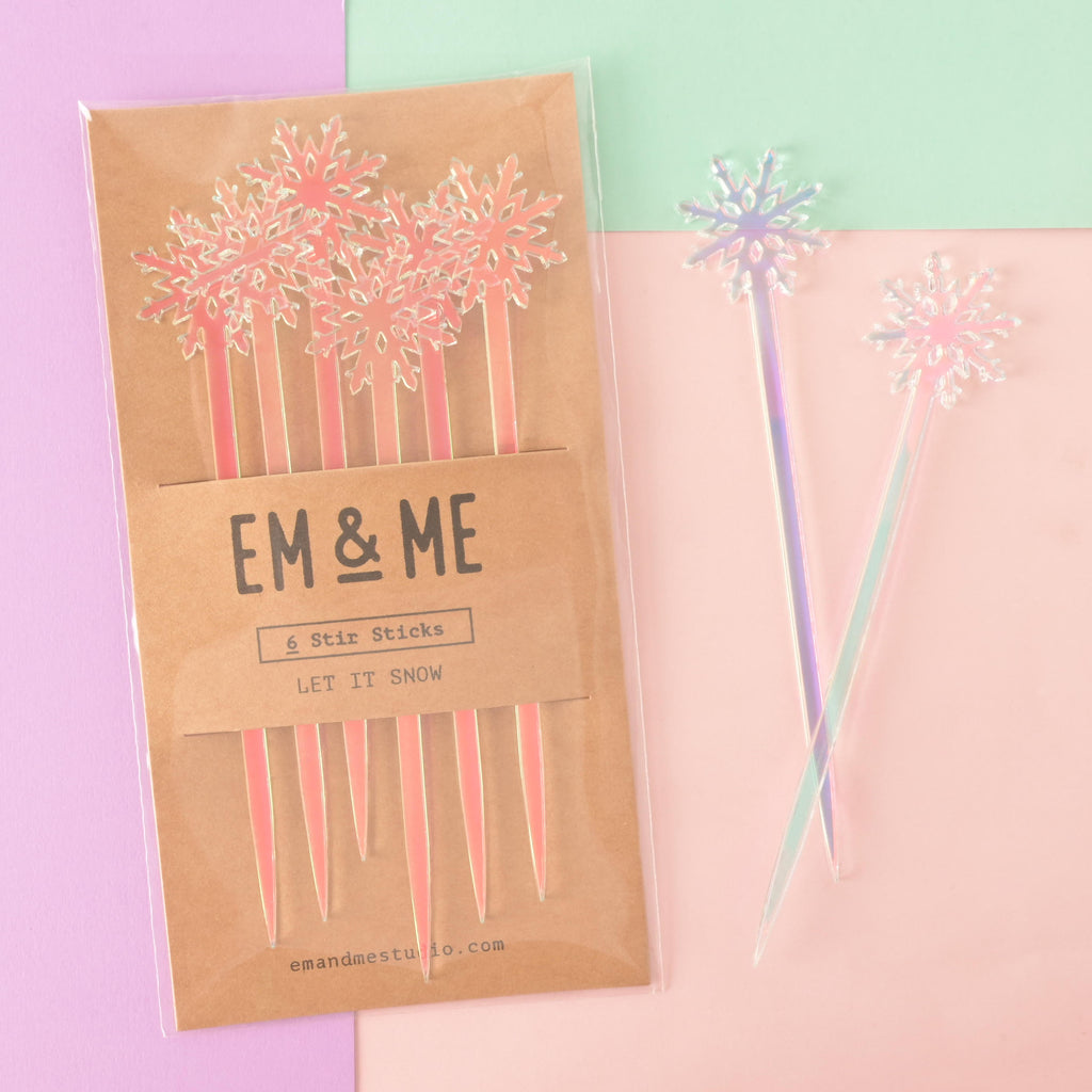 A packaged set of six pink and blue iridescent snowflake-topped cocktail stirrers. Two loose, iridescent snowflake stirrers are shown crossed next to the package. The brown card packaging reads "EM & ME" and "6 Stir Sticks" with the theme "LET IT SNOW." The items are displayed on a background split between solid pink, light purple, and mint green.