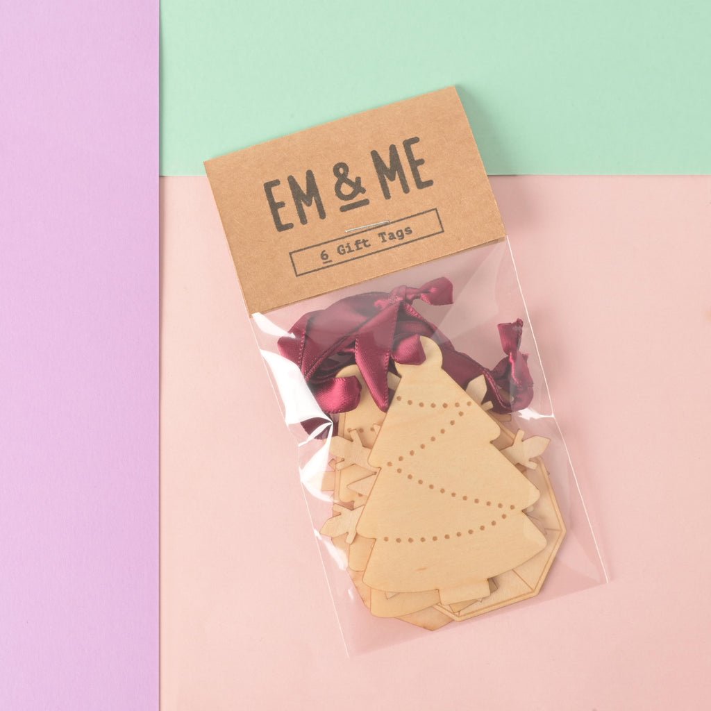 A packaged set of six blank wooden holiday gift tags in assorted shapes, including a Christmas tree with etched dots and a faceted gemstone. The packaging is a brown card that reads "EM & ME" and "6 Gift Tags." Each tag is tied with a dark red satin ribbon. The item is displayed on a background of solid pink, light purple, and mint green.