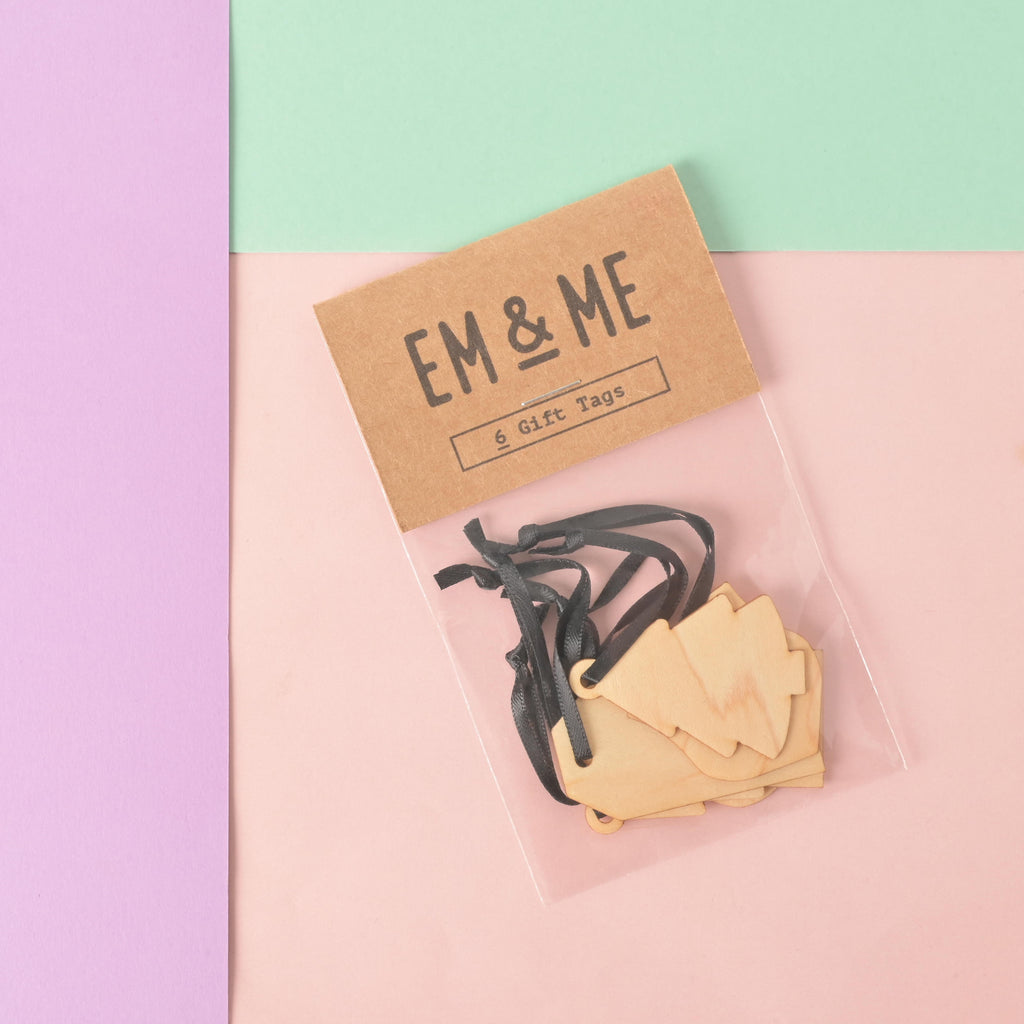 A packaged set of six blank wooden gift tags shaped like small Christmas trees. The packaging is a brown card that reads "EM & ME" and "6 Gift Tags." Each tag is tied with a small black ribbon. The item is displayed on a background of solid pink, light purple, and mint green.