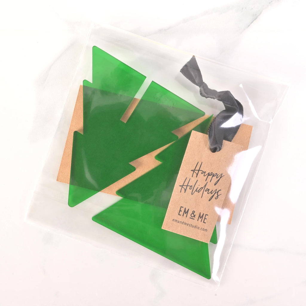 A packaged set of green flat-pack Christmas tree ornaments. The clear plastic bag contains the parts needed to assemble a tree and a brown cardstock tag that says "Happy Holidays" and "EM & ME." A dark ribbon is tied to the top of the package. The item is displayed on a white marble surface.