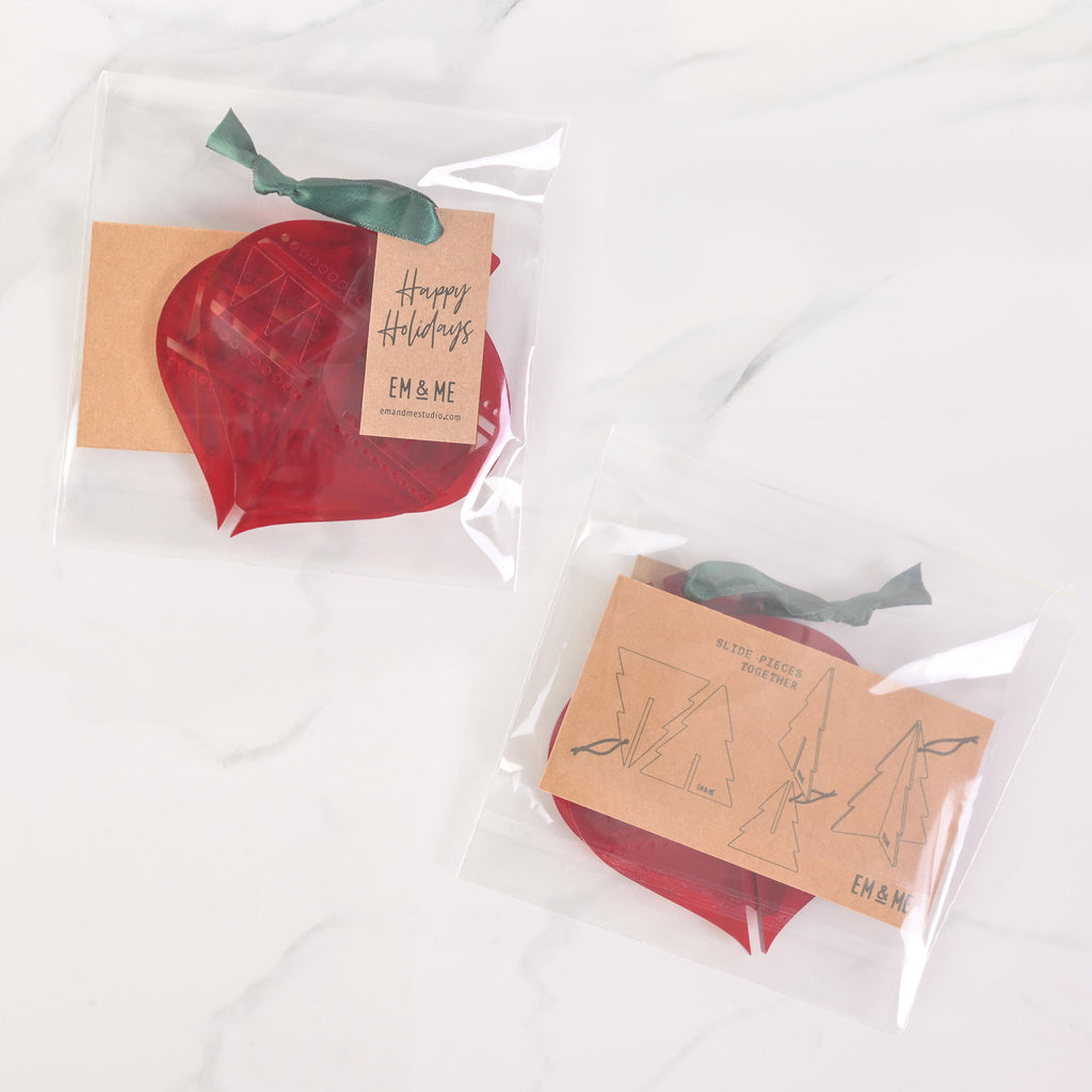 Two packaged sets of red flat-pack holiday ornaments. The package on the left contains two ornaments and has a "Happy Holidays" tag attached with a dark ribbon. The package on the right shows three ornament pieces that assemble into a Christmas tree. The packaging has "EM & ME" logos and assembly instructions. The items are displayed on a white marble surface.