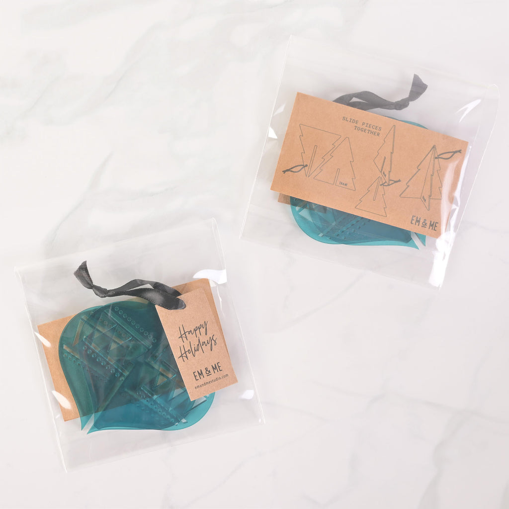 Two packaged sets of teal flat-pack holiday ornaments. The package on the left contains two ornaments and has a "Happy Holidays" tag attached with a dark ribbon. The package on the right shows three ornament pieces that assemble into a Christmas tree. The packaging has "EM & ME" logos and assembly instructions. The items are displayed on a white marble surface.