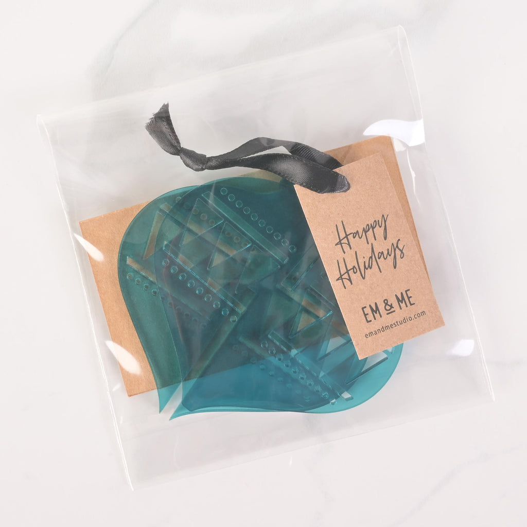 A packaged set of teal flat-pack holiday ornament pieces. The clear plastic bag contains the parts needed to assemble an ornament and a brown cardstock tag that says "Happy Holidays" and "EM & ME." A dark ribbon is tied to the top of the package. The item is displayed on a white marble surface.