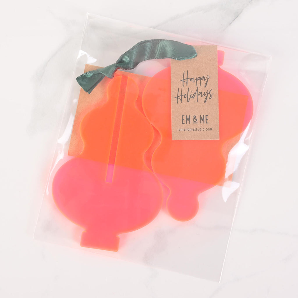 Flat Pack pink Icicle ornament The package has a "Happy Holidays" tag attached with a dark green ribbon, along with the "EM & ME" logo. The items are displayed on a white marble surface.