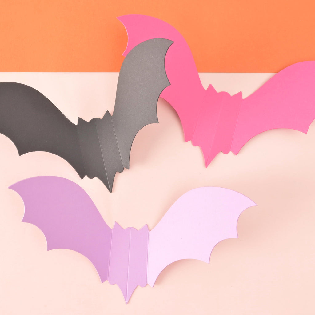 three paper bats on color blocked background showing color options of black, pink and lavender