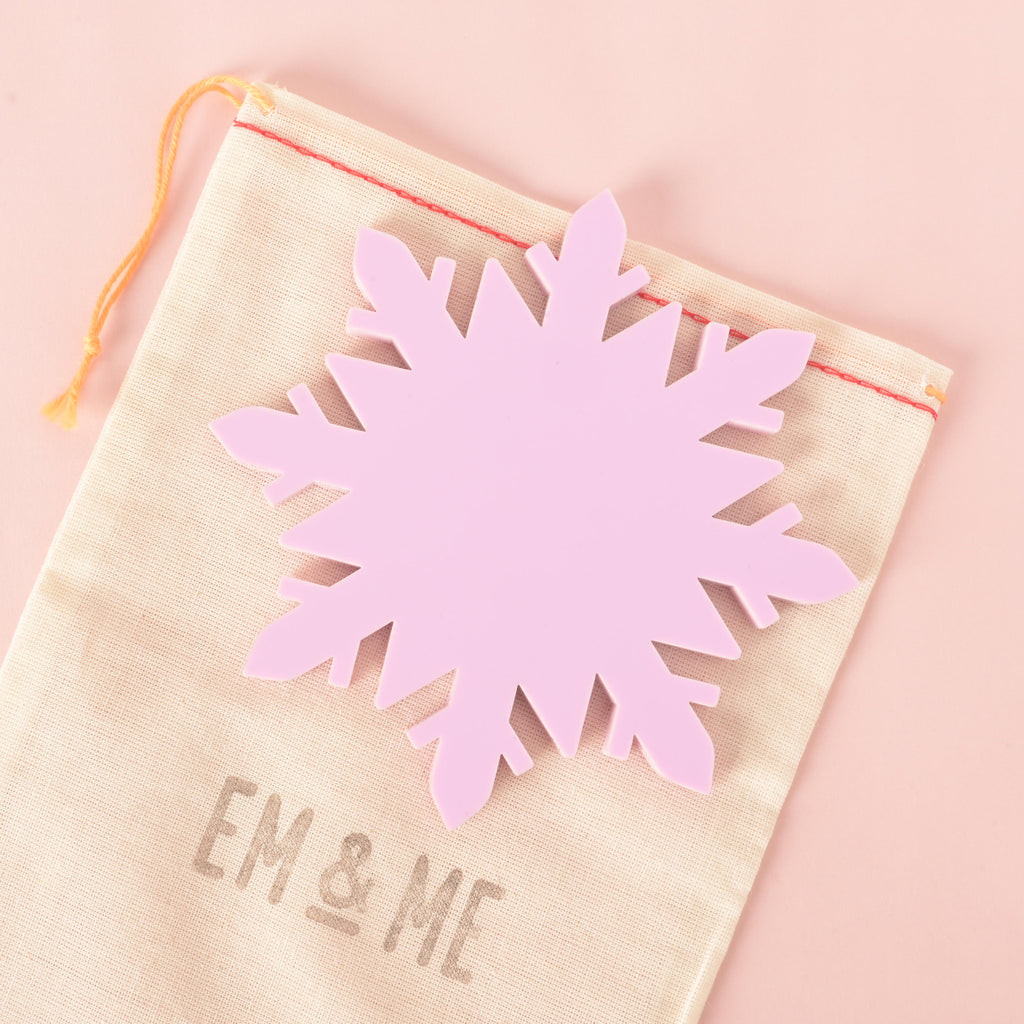 Large pastel pink snowflake-shaped Coaster from EM & ME, displayed on a beige drawstring pouch with logo, on soft pink background.
