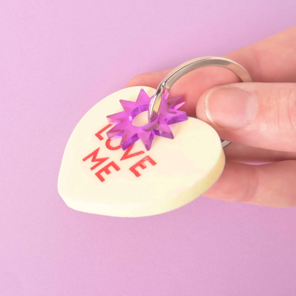 Pastel yellow Conversation Heart pet tag with red “Love Me” text, shown with purple star connector and silver keyring on lavender background.