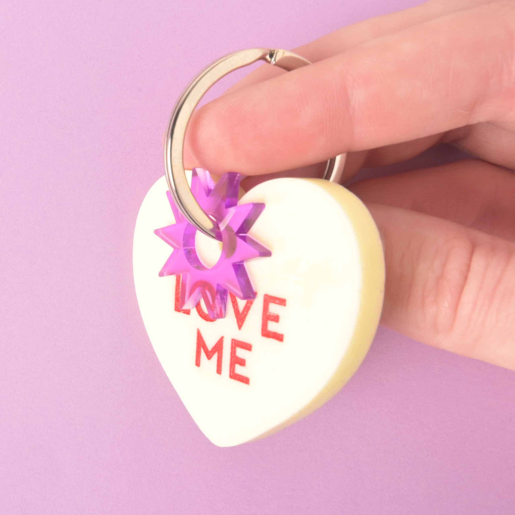 Pastel yellow heart-shaped pet ID tag with red “Love Me” text, purple star connector, and silver ring, shown held in hand on lavender backdrop.