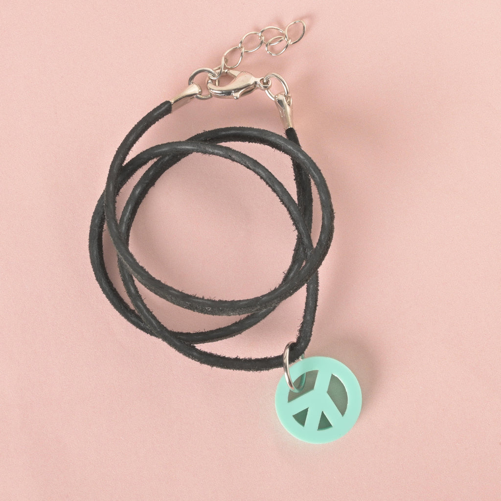A close-up, top-down view of a mint green, circular peace sign charm hanging from a black cord necklace. The necklace has a silver clasp and an extension chain. The charm and necklace are displayed on a light pink background.