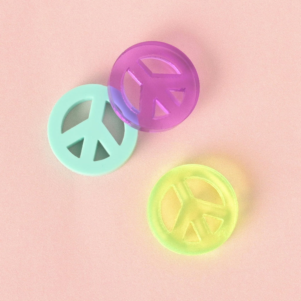 A group of three circular peace sign charms for pet tags. The charms are a translucent purple, a solid mint green, and a translucent neon green. They are arranged on a light pink background.