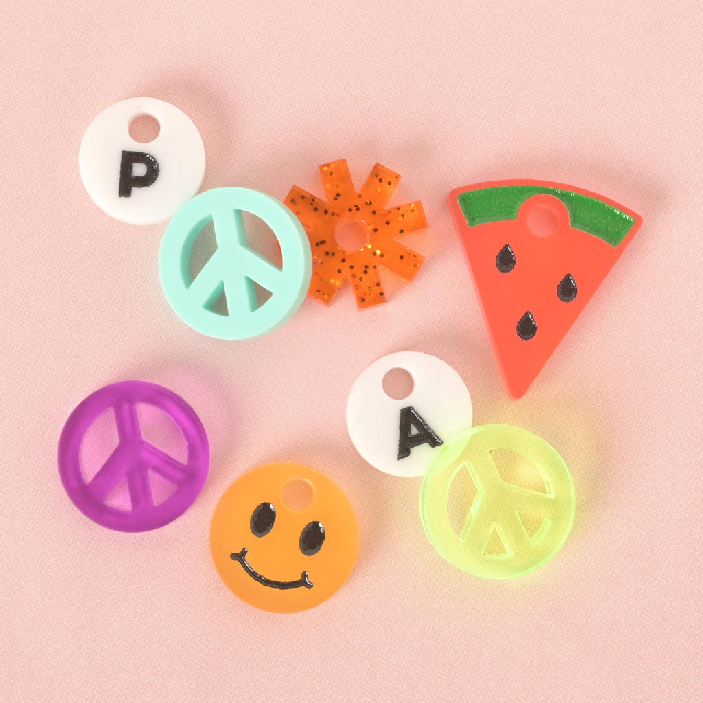 A group of colorful pet charms arranged on a pink background. The charms include two white circles with "P" and "A" printed on them, a turquoise peace sign, a translucent orange peace sign, a purple peace sign, an orange glittery sunburst, a pink and green watermelon slice, and an yellow smiley face.