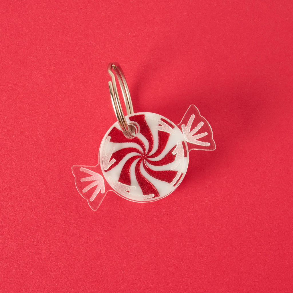 A red and white peppermint candy-shaped charm or keychain in a clear wrapper design, attached to a silver ring. The item is displayed on a solid red background.

