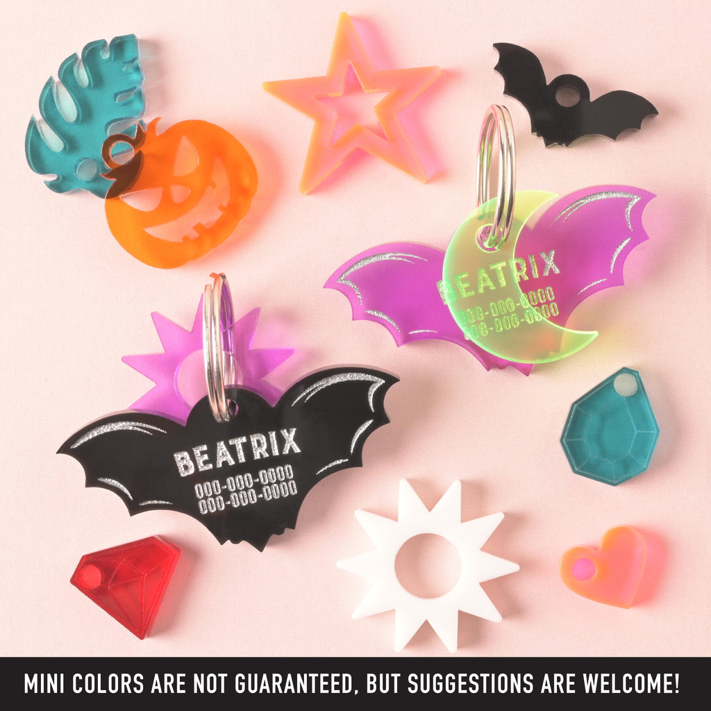 A collection of personalized acrylic bat-shaped pet ID tags for 'BEATRIX' in black and layered purple/neon green, surrounded by decorative mini charms: a turquoise leaf, an orange jack-o'-lantern, pink stars, a black bat, gem shapes, and a white sunburst.