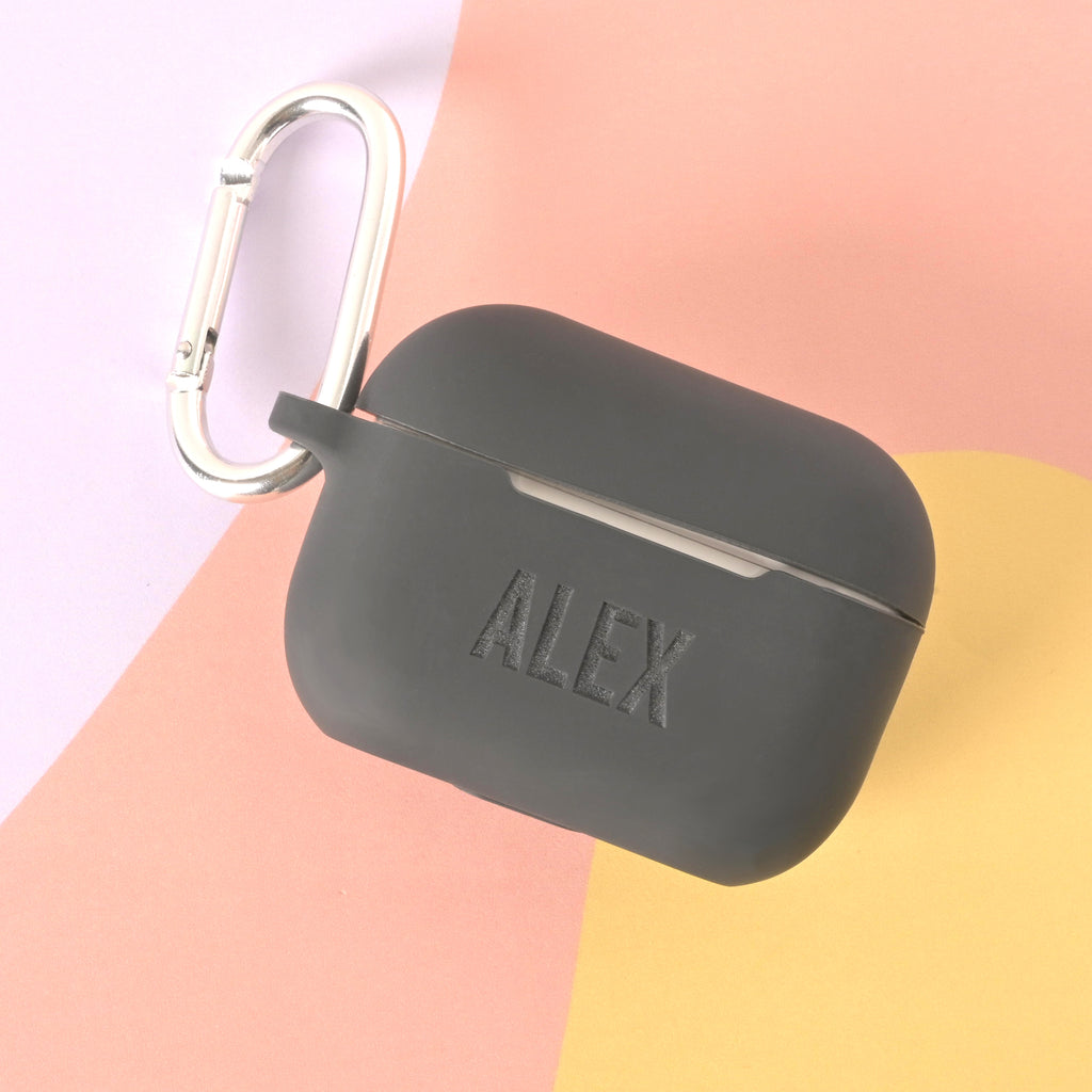 A black, custom-engraved AirPods case with a silver carabiner clip. The case is engraved with the name "ALEX" on the front. The background is a color-blocked mix of pastel pink and yellow.