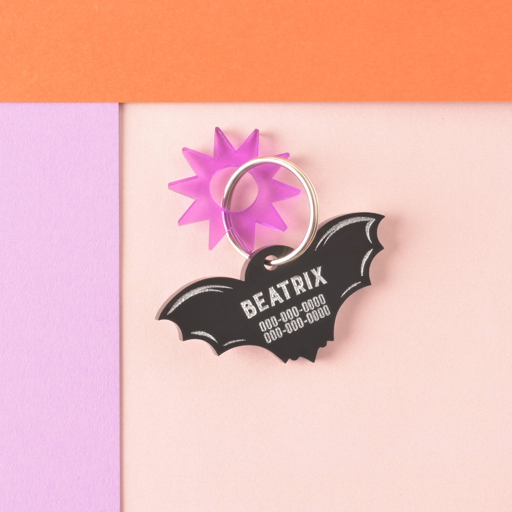 A personalized opaque black acrylic bat-shaped pet ID tag, engraved with the name 'BEATRIX' and phone numbers in white. The tag is attached to a ring with an accent charm in the shape of a translucent purple sunburst.