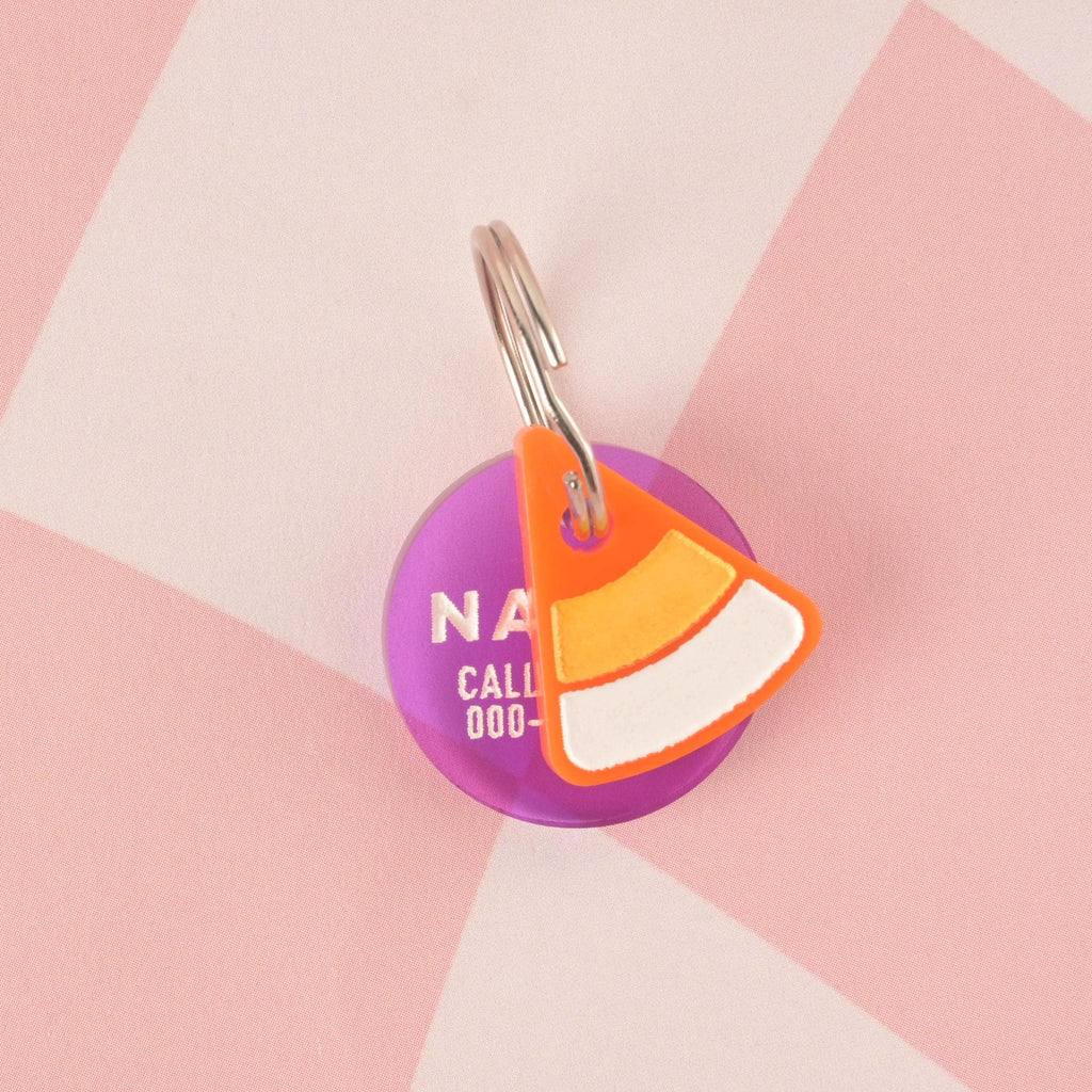 A custom pet ID tag featuring a round purple tag with text partially visible, paired with a small, layered candy corn charm. The candy corn charm has orange, yellow, and white segments and a bright orange outline. Both charms are attached to a silver keyring and are displayed on a pink and white checkered background.
