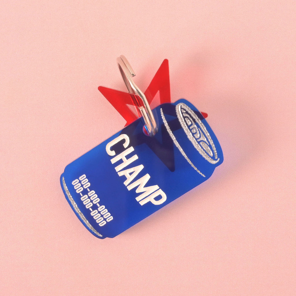 A personalized blue dog tag shaped like a soda can with the name "CHAMP" and a placeholder phone number on it. A red star charm is attached behind the tag. Both are connected to a silver ring, perfect for a collar