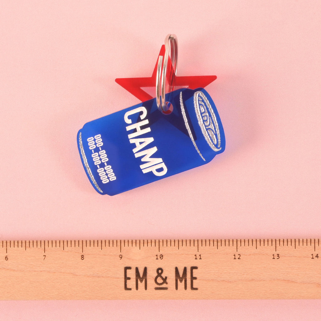 A personalized blue dog tag shaped like a soda can with the name "CHAMP" and a placeholder phone number on it. A red star charm is attached behind the tag. A wooden ruler with the "EM & ME" logo is placed below the tags to show their size. The items are displayed on a solid pink background.