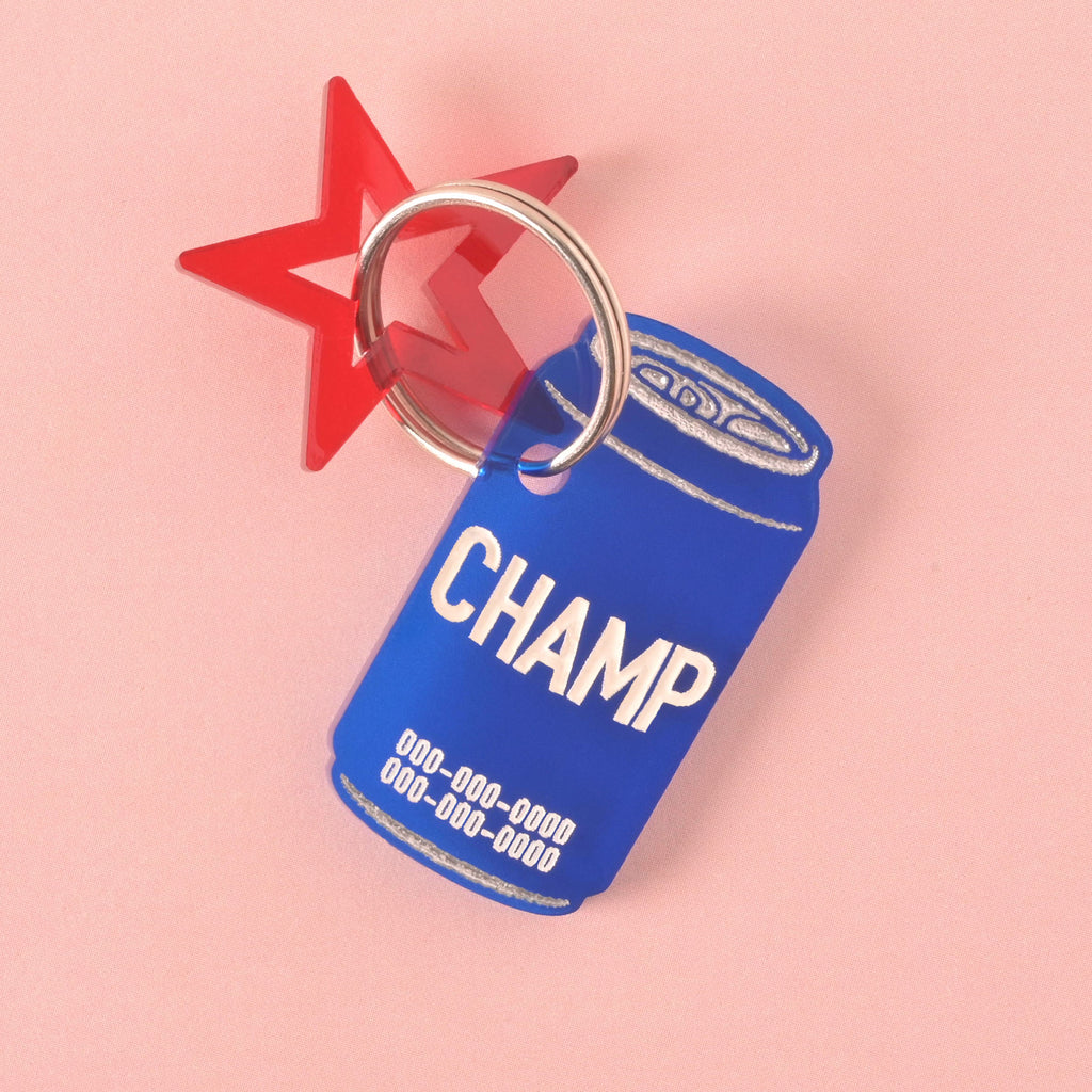 A personalized blue dog tag shaped like a soda can with the name "CHAMP" and a placeholder phone number on it. A red star charm is attached behind the tag. Both are connected to a silver ring, perfect for a collar, and are displayed on a pink background with a wavy design.