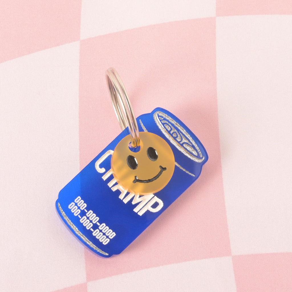 A custom, blue, can-shaped pet ID tag with "CHAMP" engraved on it. The can tag is also engraved with a phone number in white font. A small, translucent yellow smiley face charm is attached to the silver keyring. The tags are displayed on a pink and white checkered background.