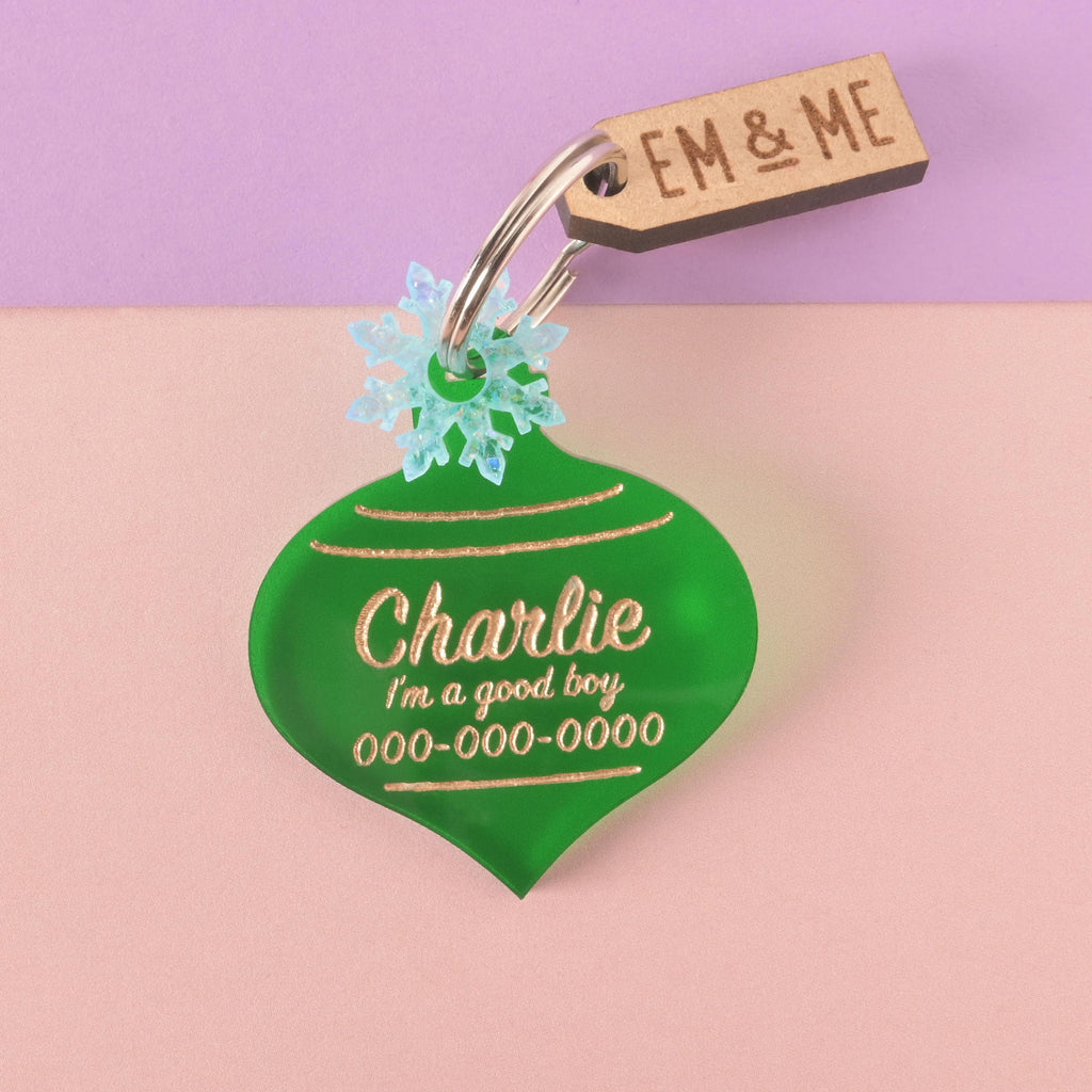 A personalized green ornament-shaped pet tag with the name "Charlie," the phrase "I'm a good boy," and a placeholder phone number on it. The tag has a small, light blue iridescent snowflake charm and a wooden "EM & ME" tag attached. Both are connected to a silver ring, perfect for a collar, and are displayed on a background that is half solid purple and half solid pink.