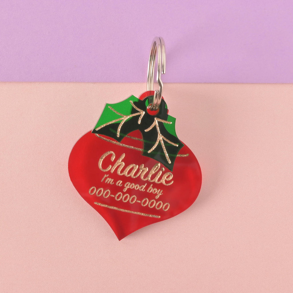 A personalized red ornament-shaped pet tag with the name "Charlie," the phrase "I'm a good boy," and a placeholder phone number on it. The tag is decorated with green holly leaves. It's attached to a silver ring, perfect for a collar, and is displayed on a background that is half solid purple and half solid pink.