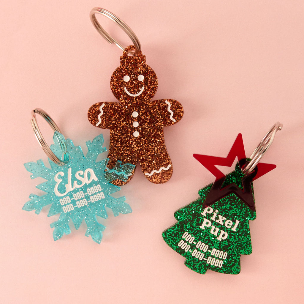 A collection of three custom, glitter acrylic holiday pet ID tags: a blue snowflake tag for 'Elsa', a brown gingerbread man charm, and a green Christmas tree tag for 'Pixel Pup' with a red star accent. All tags are personalized with names and contact information.