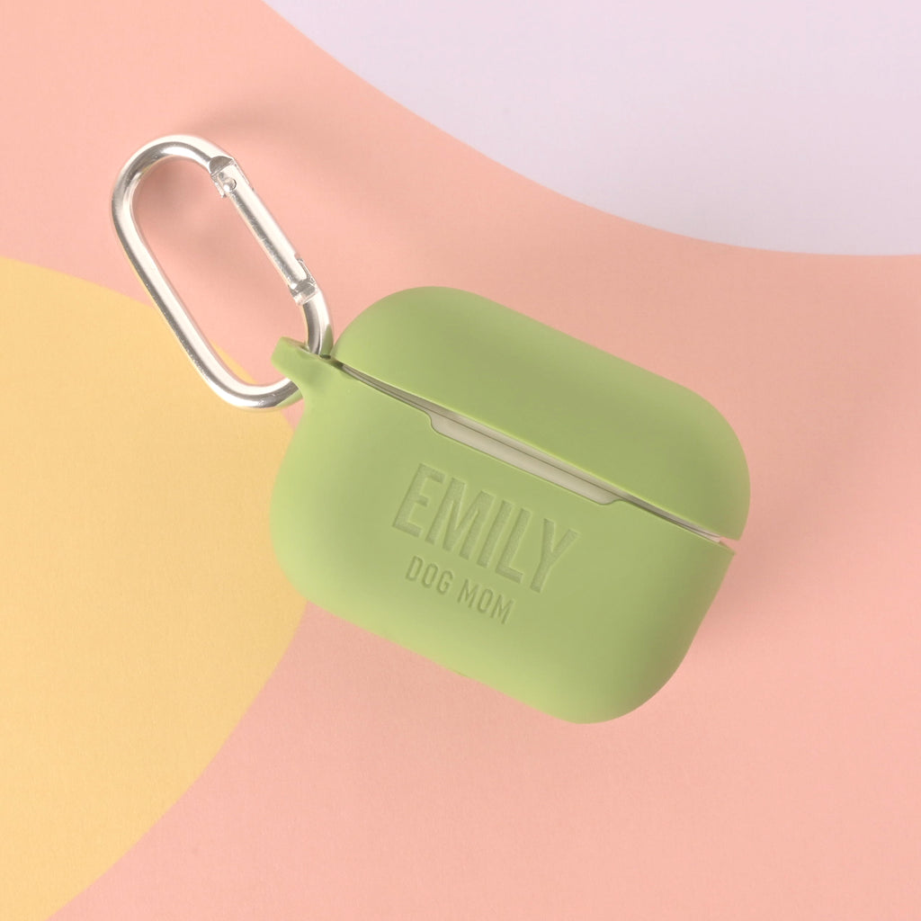 A custom sage green AirPods case with a silver carabiner clip. The case is engraved with "EMILY" in large letters and "DOG MOM" below. The background is pastel pink, purple, and yellow.