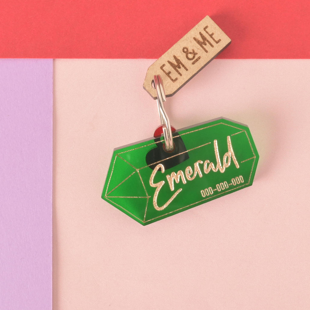A personalized green gemstone-shaped pet tag with the name "Emerald" and a placeholder phone number on it. A small black heart charm and a wooden "EM & ME" tag are attached. Both are connected to a silver ring for a collar and are displayed on a light pink, purple, and red background.