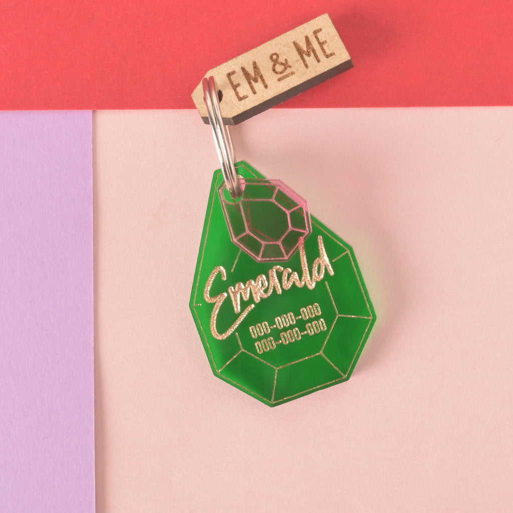 A personalized green gemstone-shaped pet tag with the name "Emerald" written in a gold-colored script, along with a placeholder phone number. A small pink gemstone charm is attached to the tag, along with a small wooden charm that says "EM & ME." All are connected to a silver ring for a collar and are displayed on a light pink, purple, and red background.