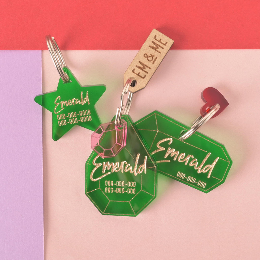 A collection of three personalized green pet tags with the name "Emerald" and a placeholder phone number on each. The tags include a star-shaped one, a large gemstone-shaped one with a pink gemstone charm, and a rectangular one with a small red heart charm. A wooden "EM & ME" tag is also visible. All items are attached to a silver ring for a collar and are displayed on a light pink, purple, and red background.