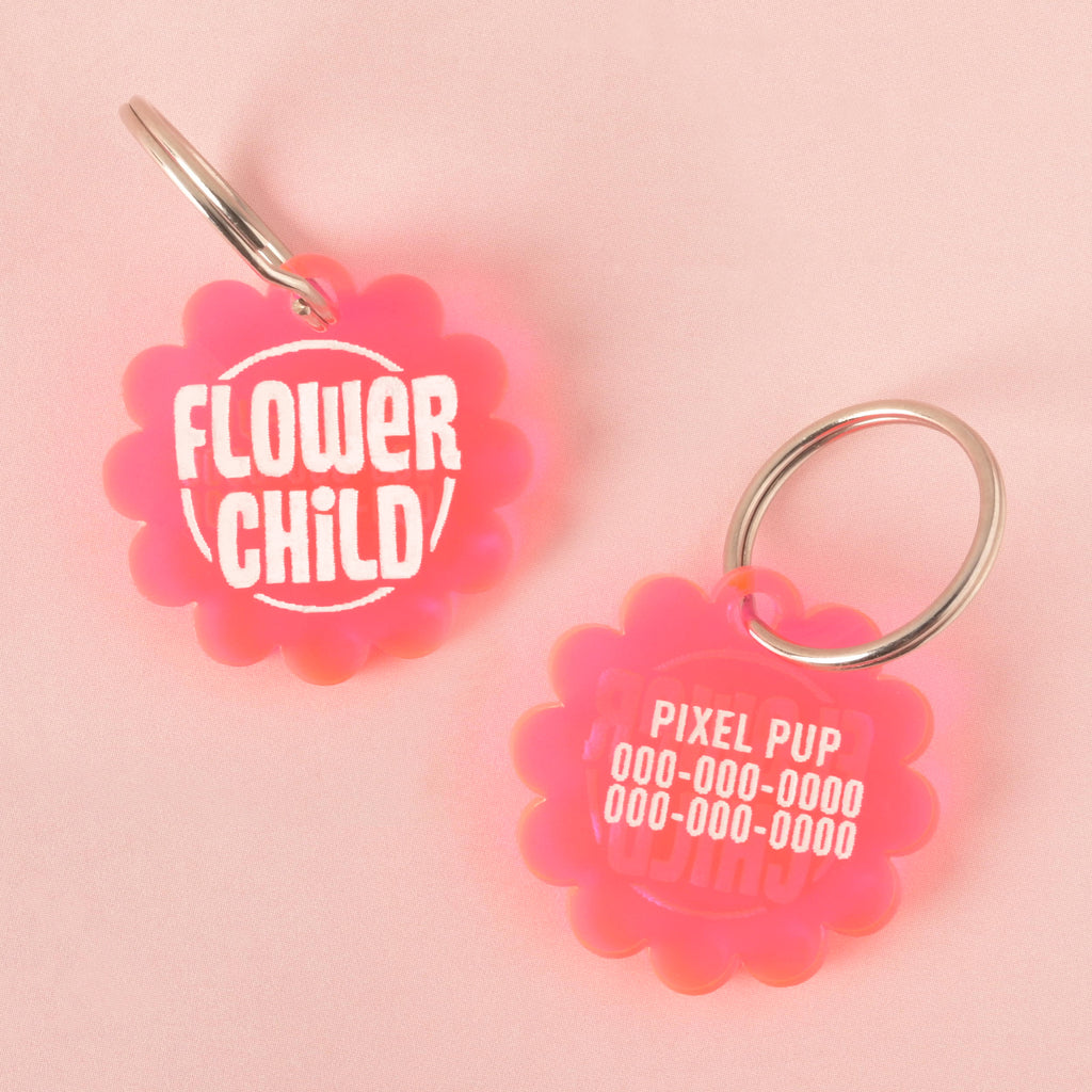 A pair of pink flower-shaped pet tags. The front of one tag says "FLOWER CHILD," while the back of the other has a placeholder for a pet's name, "PIXEL PUP," and a phone number. Both tags are attached to a silver ring for a collar and are displayed on a light pink background.