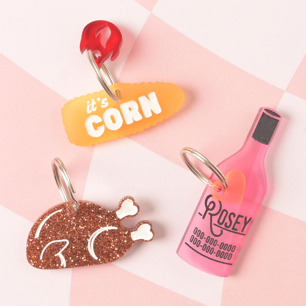 A collection of four personalized pet tags with food-themed designs. The tags include a pink wine bottle for "ROSEY," a glittery turkey leg, a yellow corn on the cob that says "it's CORN," and a small red charm. All tags are attached to silver rings and are displayed on a pink and white checkered background.