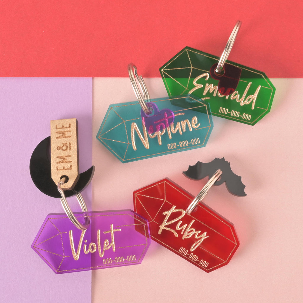 A collection of four personalized gemstone-shaped pet tags in various colors on a background of light pink, purple, and red. The tags include a purple gemstone for "Violet," a translucent teal gemstone for "Neptune," a red gemstone for "Ruby," and a green gemstone for "Emerald." Each tag has a placeholder for a phone number and is attached to a silver ring for a collar. Other charms like a black moon, a black bat, and a small wooden "EM & ME" tag are also visible.