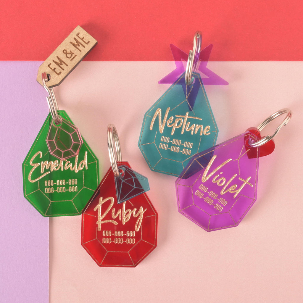 A collection of four personalized gemstone-shaped pet tags in various colors on a background of light pink, purple, and red. The tags include a green gemstone for "Emerald," a red gemstone for "Ruby," a teal gemstone for "Neptune," and a purple gemstone for "Violet." Each tag has a placeholder phone number and is attached to a silver ring for a collar. The items are shown with small charms like a wooden "EM & ME" tag, a pink gemstone, a purple starburst, and a red heart.