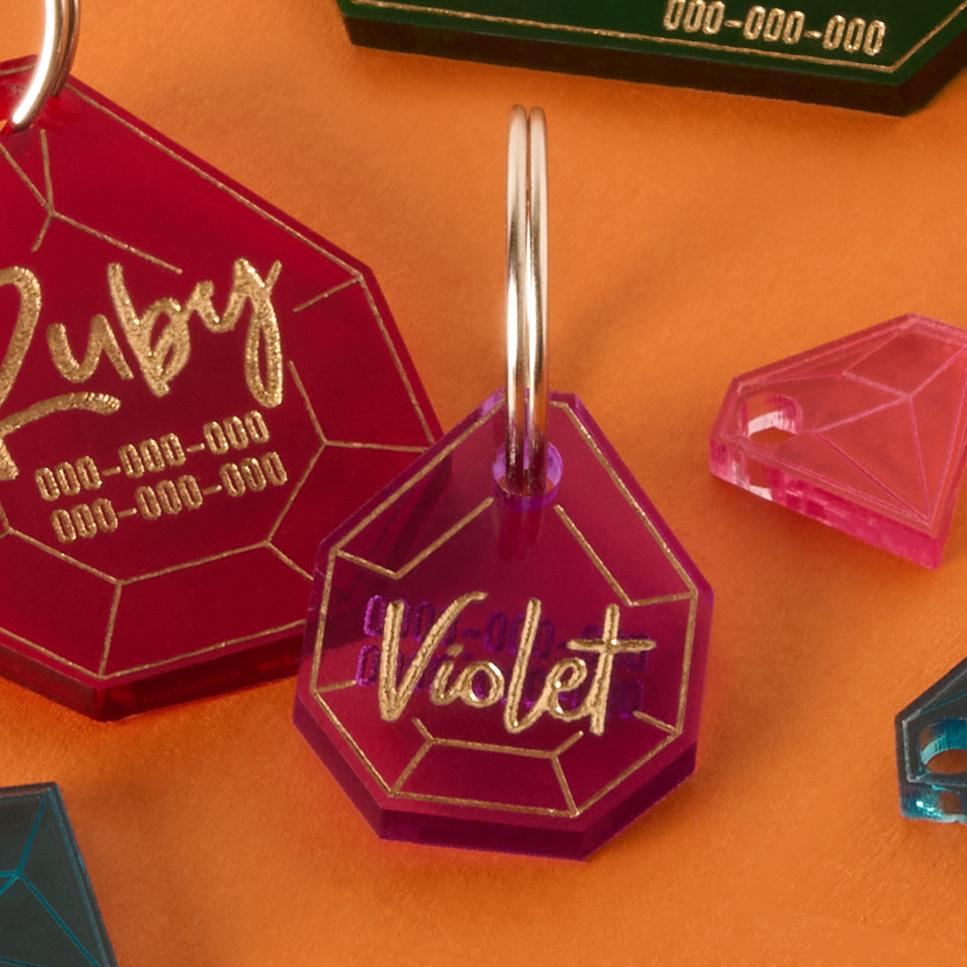 A close-up of two personalized, gemstone-shaped pet tags on a background that is half solid orange and half solid pink. The larger red tag has the name "Ruby" and a placeholder phone number. The smaller purple tag has the name "Violet" written in a gold-colored script. Both tags are attached to a silver ring for a collar.
