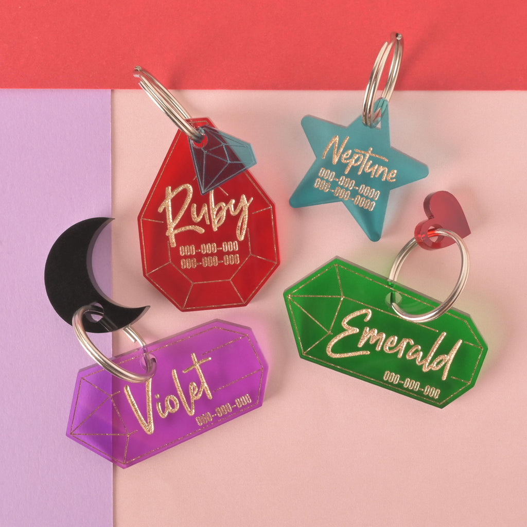 A collection of four personalized gemstone and star-shaped pet tags in various colors on a background of light pink, purple, and red. The tags include a red gemstone for "Ruby," a teal star for "Neptune," a purple gemstone for "Violet," and a green gemstone for "Emerald." Each tag has a placeholder phone number and is attached to a silver ring for a collar. Small charms like a black moon and a red heart are also visible on the tags.