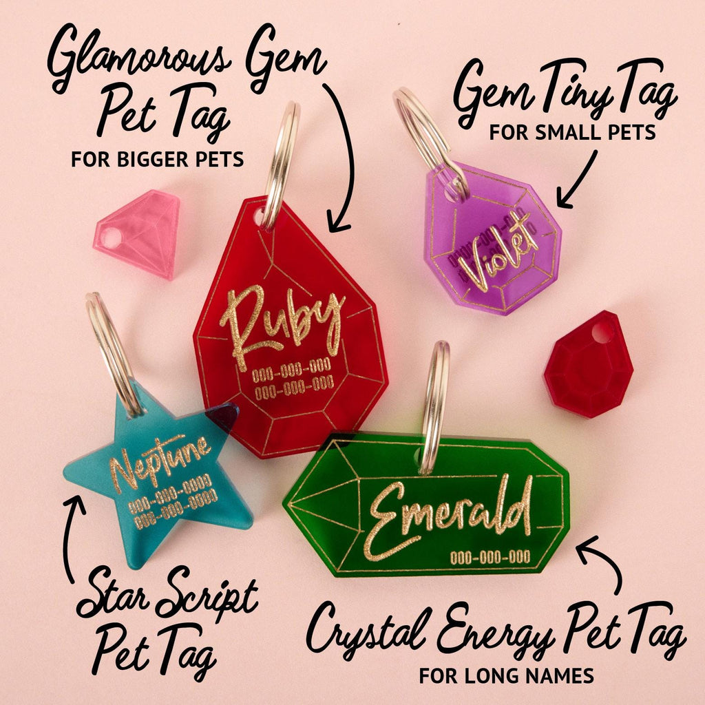 A collection of personalized pet tags in various gemstone and star shapes. The tags include a large red gemstone that says "Ruby," a purple gemstone for "Violet," a teal star for "Neptune," and a green gemstone for "Emerald." The tags are shown with text describing their sizes and designs, such as "Glamorous Gem Pet Tag for Bigger Pets" and "Gem Tiny Tag for Small Pets." The items are displayed on a solid pink background.
