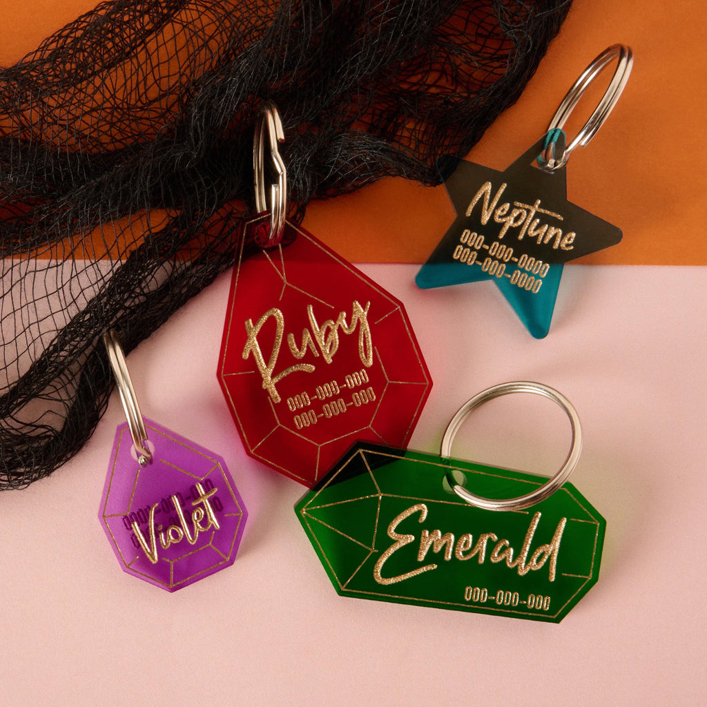 A collection of four personalized pet tags in various shapes and colors on a background of black netting and a split surface of orange and pink. The tags include a large red gemstone that says "Ruby," a purple gemstone for "Violet," a teal star for "Neptune," and a green gemstone for "Emerald." Each tag has a placeholder for a phone number and is attached to a silver ring for a collar.