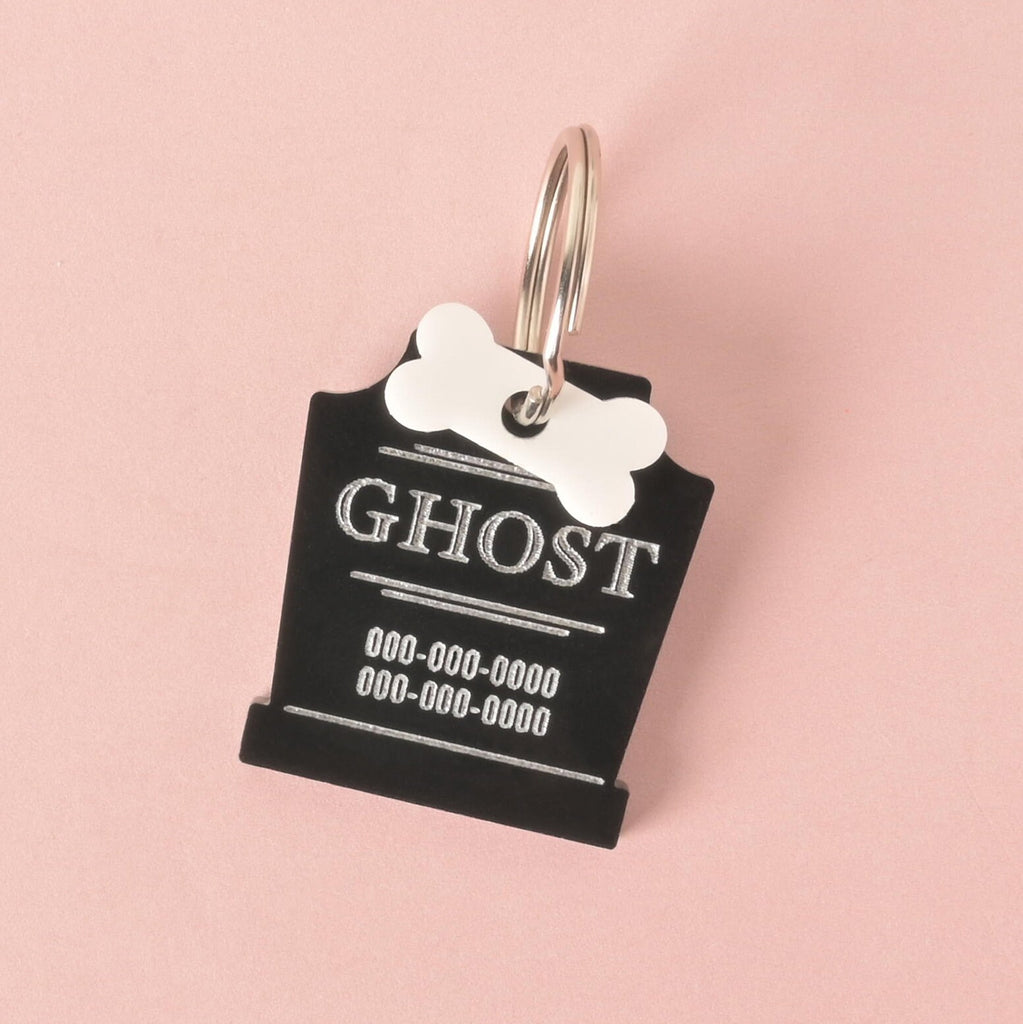 A close-up shot of a custom, black, gravestone-shaped pet ID tag with a shiny silver keyring. The tag is engraved with the name "GHOST" and a phone number in white font. A small white bone charm is attached, sitting on top of the gravestone shape. The tag is displayed on a solid pink background.
