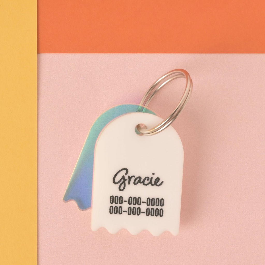 A custom, white ghost-shaped pet ID tag with a wavy bottom. The tag is engraved with the name "Gracie" in script and a phone number below it. A small, iridescent ghost charm hangs behind the main tag. Both are attached to a silver keyring and are set against a pink, orange, and yellow color-blocked background.