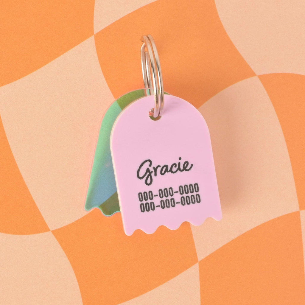 A custom, Lavender ghost-shaped pet ID tag with a wavy bottom. The tag is engraved with the name "Gracie" in script and a phone number below it. A second, translucent green and blue iridescent ghost charm is visible behind it. Both are attached to a silver keyring and are displayed on a checkered orange and beige background.