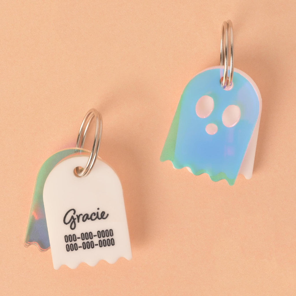 Two custom ghost-shaped pet ID tags on a peach-colored background. The tag on the left is a white ghost with "Gracie" and a phone number engraved on it, and a translucent iridescent charm behind it. The tag on the right is a translucent iridescent ghost with a phone number engraved on it. Both have silver keyrings.