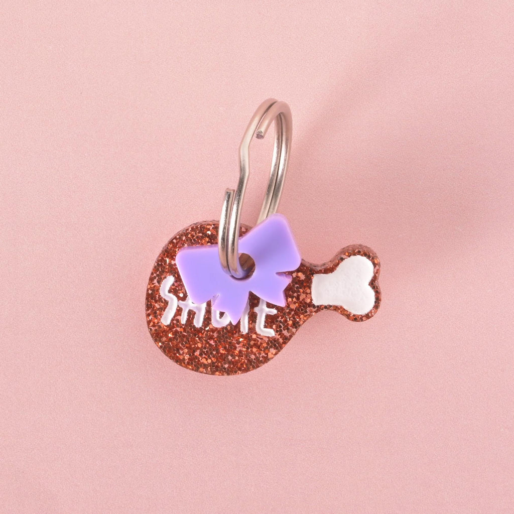 Small glittery drumstick-shaped pet ID tag with the name “Sadie” in white letters, decorated with a purple bow charm and silver key ring.