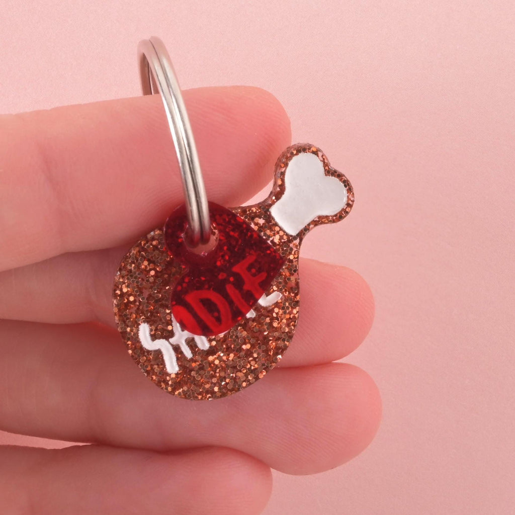 Close-up of a glittery drumstick-shaped pet ID tag with the name “Sadie” in white letters, attached to a silver key ring, held between fingers.