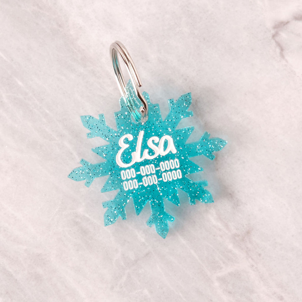A custom, laser-cut pet ID tag shaped like a snowflake in glittery blue acrylic. The tag is engraved with the name 'Elsa' and three lines of contact information in white, set against a light marble background.