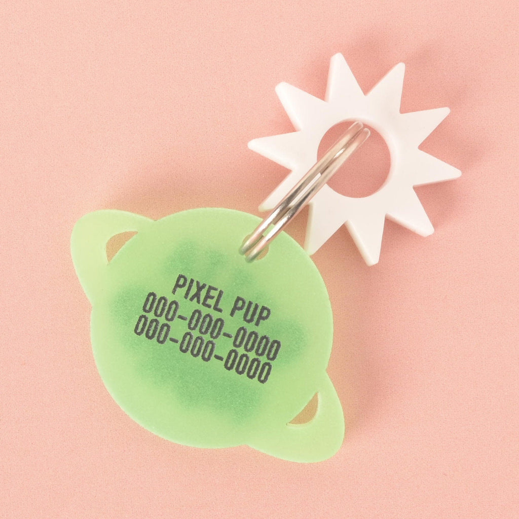 A custom, glow-in-the-dark, planet-shaped pet ID tag with a silver keyring. The tag is engraved with the text "PIXEL PUP" and a phone number. A small white sunburst charm is attached to the keyring. The tag is shown on a solid pink background.