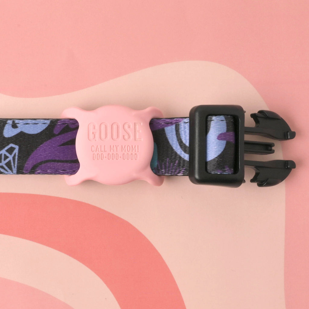 A custom, pink, silent pet ID tag with "GOOSE," "CALL MY MOM!", and a phone number engraved on it. The tag is shown slid onto a black and purple patterned pet collar with a black buckle. The collar is set against a light pink and reddish-brown wavy-patterned background.